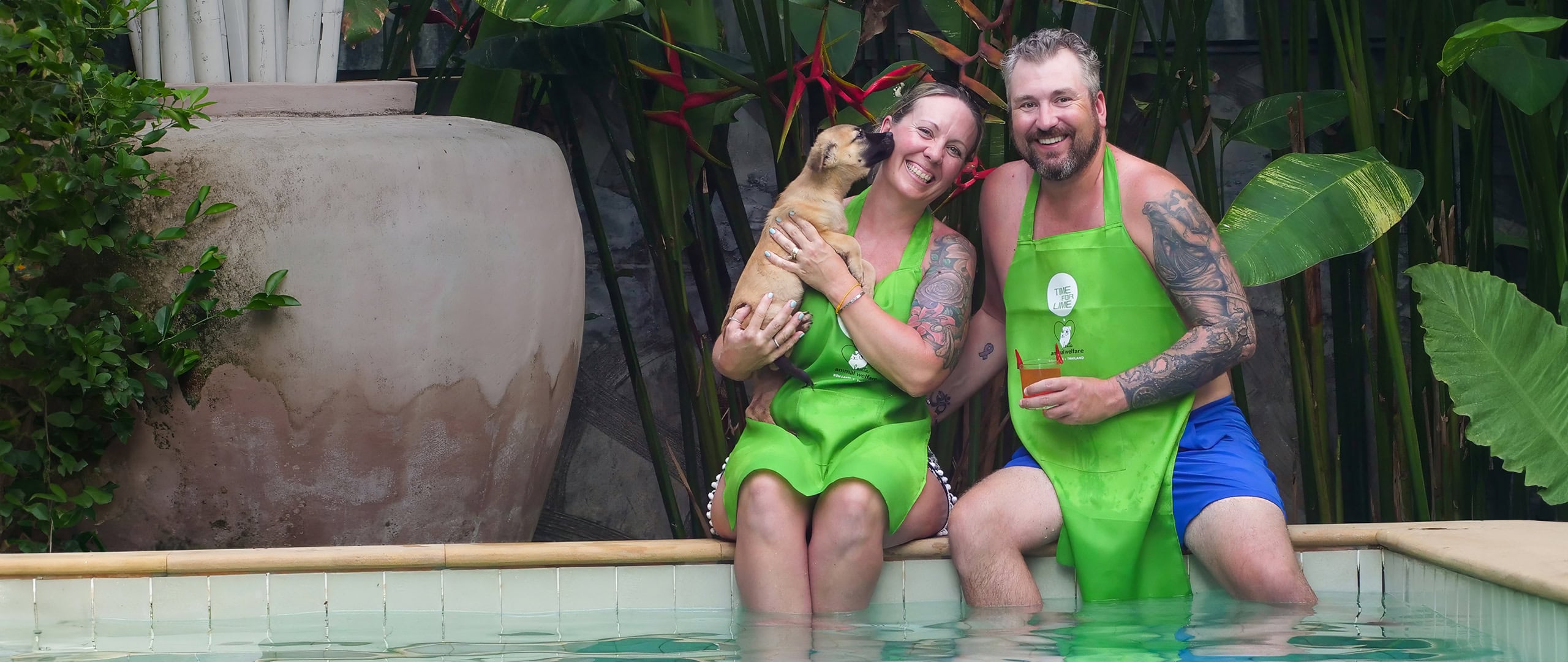 A smiling couple in bright green aprons sits by a pool, with a playful puppy licking the woman’s face. The man holds a drink, and tropical plants surround them.